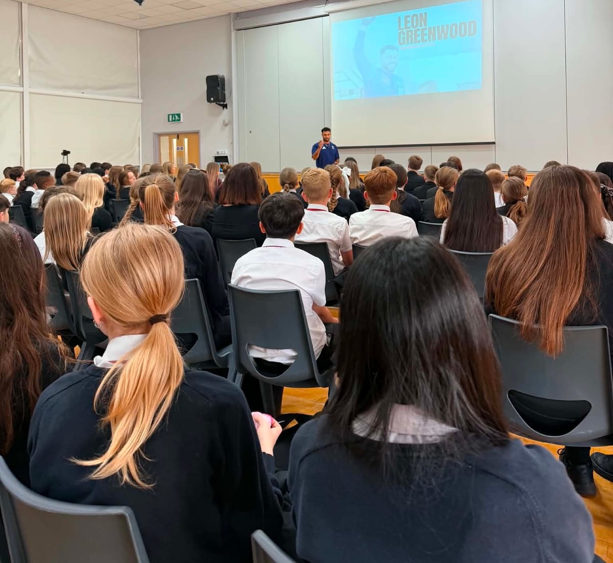 Leon speaks to a packed hall of secondary students, seen from among the audience, with his name on the screen behind him.