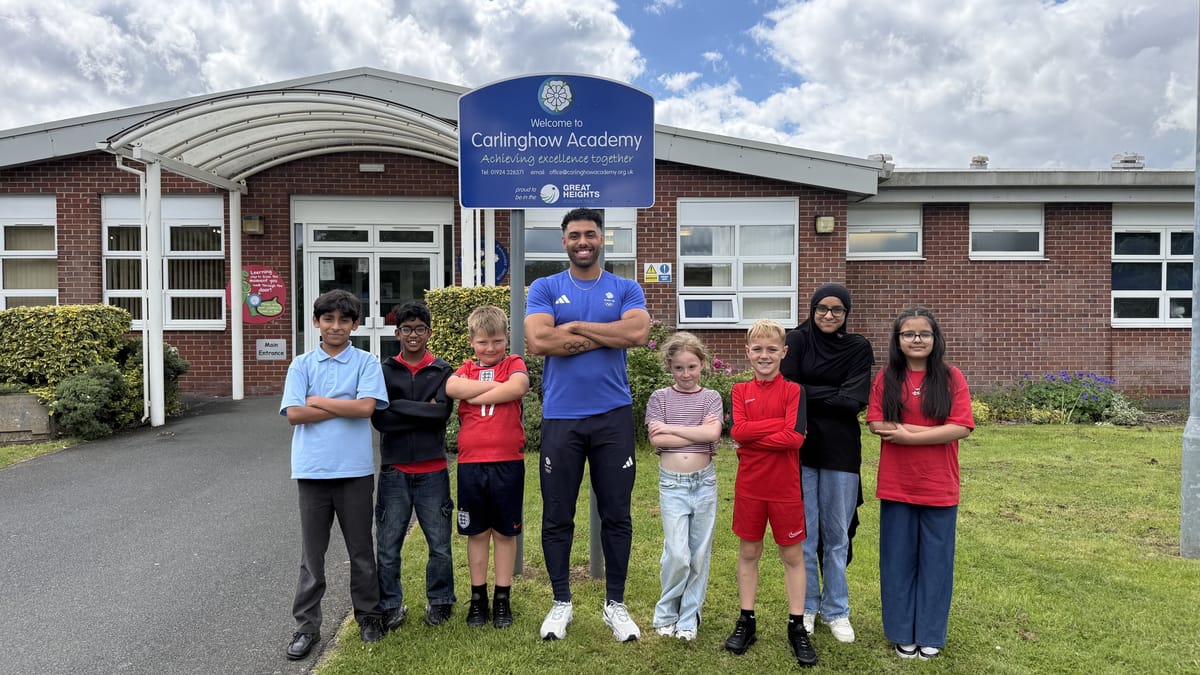 Leon Greenwood stands with arms folded alongside a group of pupils outside their school on a sunny day.