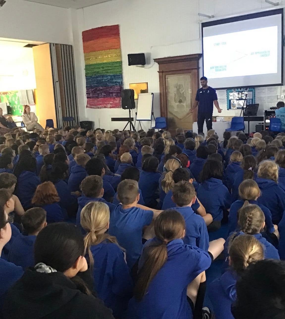 Rows of primary pupils in blue jumpers sit on a hall floor facing a speaker and a projected slide.