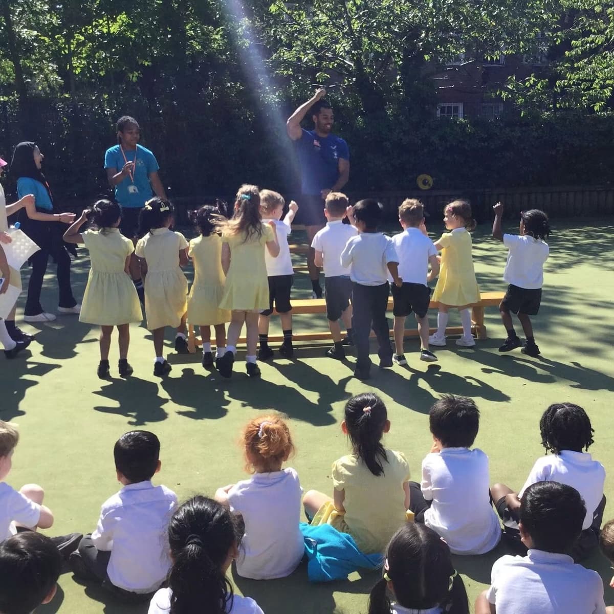 Primary pupils in yellow and white uniforms dance and cheer in a sunny playground while their classmates watch, sitting on the ground.